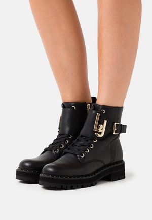 Bottes chevilles en cuir noir avec devant à lacets, sangles à boucle en métal doré, et semelles épaisses crantées portées sur des jambes nues.