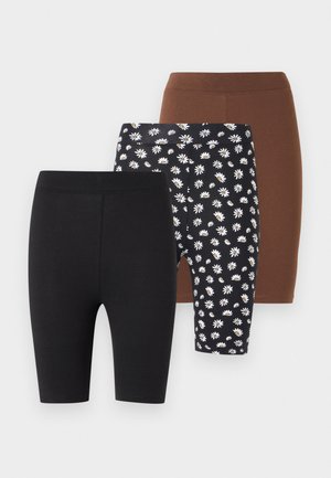 Jeune femme debout sur fond blanc portant un crop top rose pâle à encolure carrée, un short noir ajusté longueur genou et des tongs noires.
