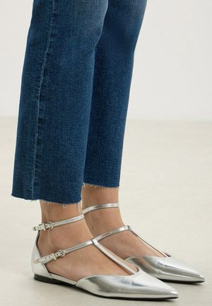 Feet wearing silver pointed-toe flats with ankle and foot straps, paired with cropped dark blue jeans with raw hems.