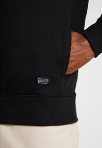 Sudadera negra con un acabado texturizado, que presenta un bolsillo lateral y una etiqueta azul en el dobladillo. Puños y cintura acanalados.