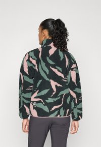 Donna con coda di cavallo riccia che indossa una giacca in pile a fantasia nera, rosa e verde e pantaloni grigi, di spalle contro uno sfondo neutro.