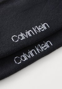 Fekete pamut zoknik, amelyekhez egy fehér "Calvin Klein" logó van hímzve a szárnál, sima textúrával és testre szabott kialakítással.