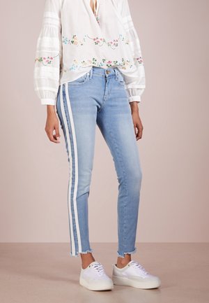 Personne portant un jean skinny bleu clair avec des bandes blanches sur les côtés, un chemisier blanc brodé de fleurs, et des baskets blanches, debout sur un sol neutre.