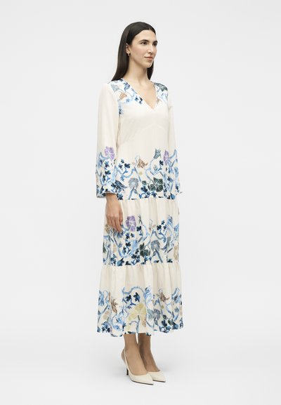 Femme aux longs cheveux foncés portant une robe longue crème à manches longues avec une broderie florale bleue, violette et beige, debout en talons blancs.