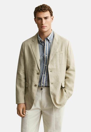 Jonge man die een beige blazer draagt over een blauw-wit gestreept overhemd met knopen en een beige broek, staand met één hand in de zak.