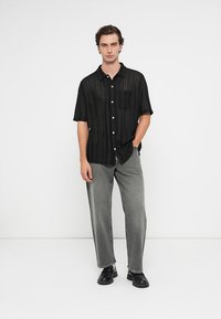 Vyras stovi vilkėdamas juodą marškinėlių trumpomis rankovėmis mygtuką, pilkas džinsus ir juodus batus prieš baltą foną.