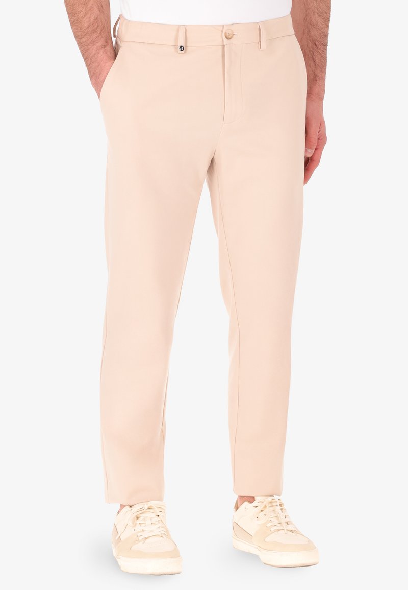 Pantaloni beige con vestibilità slim, realizzati in tessuto morbido, con tasche laterali e chiusura con bottone. Abbinati a sneakers di colore chiaro.