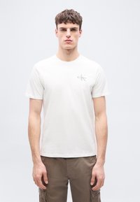 Νέος άνδρας που στέκεται προς τα εμπρός, φορώντας ένα απλό λευκό T-shirt Calvin Klein και καφέ παντελόνι cargo σε φωτεινό φόντο.