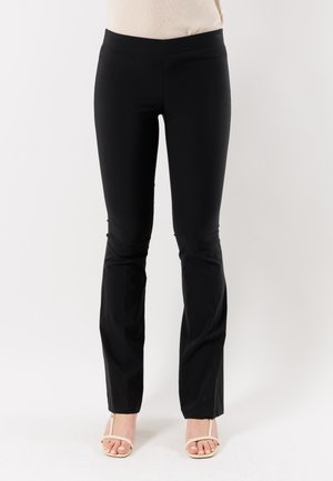 Donna con pantaloni neri aderenti e tacchi beige aperti in punta, in piedi davanti a uno sfondo bianco neutro.