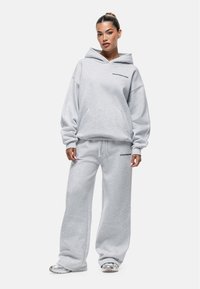 Jonge vrouw met een hoge paardenstaart, gekleed in een lichtgrijze hoodie en bijpassende wijde sweatpants met kleine zwarte tekst op de borst en bovenbeen.
