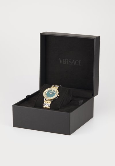 Montre dorée avec un cadran turquoise, une lunette à motifs et un bracelet accentué de pyramides, présentée dans une boîte noire de Versace avec un intérieur doux.
