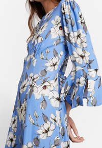 Blue floral dress featuring large white flowers with gray leaves. The fabric has a soft texture, with bell sleeves and cut-out details.