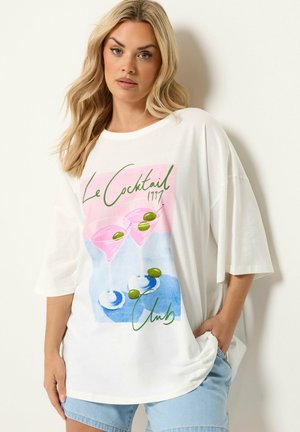 Blonde Frau trägt weißes oversized T-Shirt mit pinken Cocktailgläsern und grünen Oliven als Grafik, kombiniert mit hellblauen Jeansshorts.