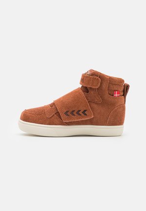 Scarpa alta in suede marrone con strap in Velcro, suola bianca e dettagli neri a zig-zag. Il lato presenta un piccolo dettaglio a forma di bandiera.