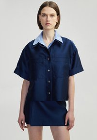 Navy blue satin short-sleeve shirt with a light blue collar, front button closure, and two chest pockets. Styled with a matching skirt.