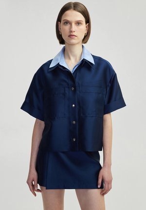 Navy blue satin short-sleeve shirt with a light blue collar, front button closure, and two chest pockets. Styled with a matching skirt.