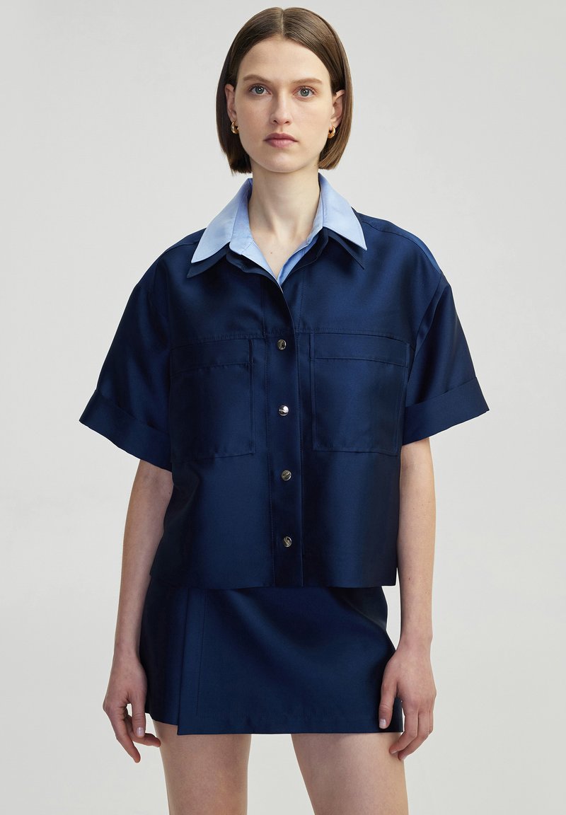 Navy blue satin short-sleeve shirt with a light blue collar, front button closure, and two chest pockets. Styled with a matching skirt.