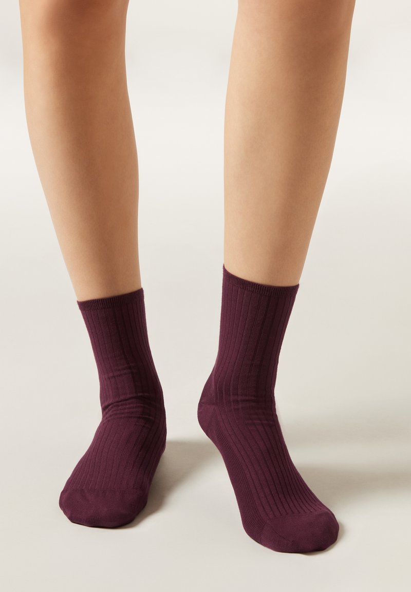 Jambes portant des chaussettes côtelées bordeaux mi-mollet, debout sur un sol clair et neutre.