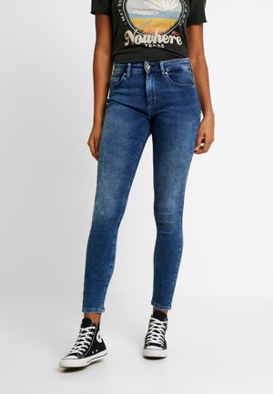 Osoba ubrana w ciemnoniebieskie obcisłe dżinsy, czarne wysokie trampki oraz czarną koszulkę z grafiką, stojąca na tle jednolitego tła.