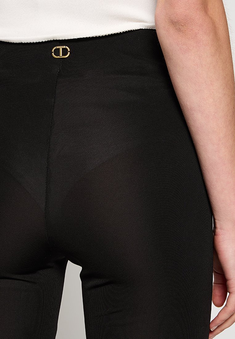 Leggings negros hechos de una tela suave y elástica. Cuenta con un detalle de logo dorado en la cintura. El diseño es ajustado y elegante.
