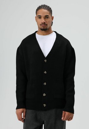 Zwarte gebreide cardigan met een V-hals, zes metalen knopen, twee voorzakken en geribde manchetten en zoom. Gedragen over een wit overhemd.