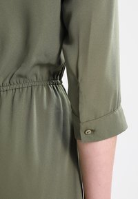 Oliwkowa sukienka z gładką, błyszczącą teksturą. Posiada ściągacz w pasie oraz guziki na mankietach rękawów o długości trzech czwartych.