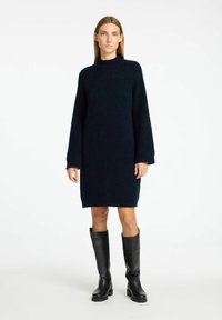 Tumši zils adīts kleita ar garām piedurknēm un augstu kaklu, izgatavota no mīkstas, teksturētas auduma, kombinēta ar melnām ādas zābakiem līdz ceļgaliem.