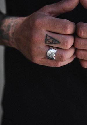 Tekstuuriga hõbesõrmus ebaühtlase pinnaga, kantud geomeetrilise tätoveeringuga käele. Käsi on lõdvestunud asendis, ring paistab hästi silma.