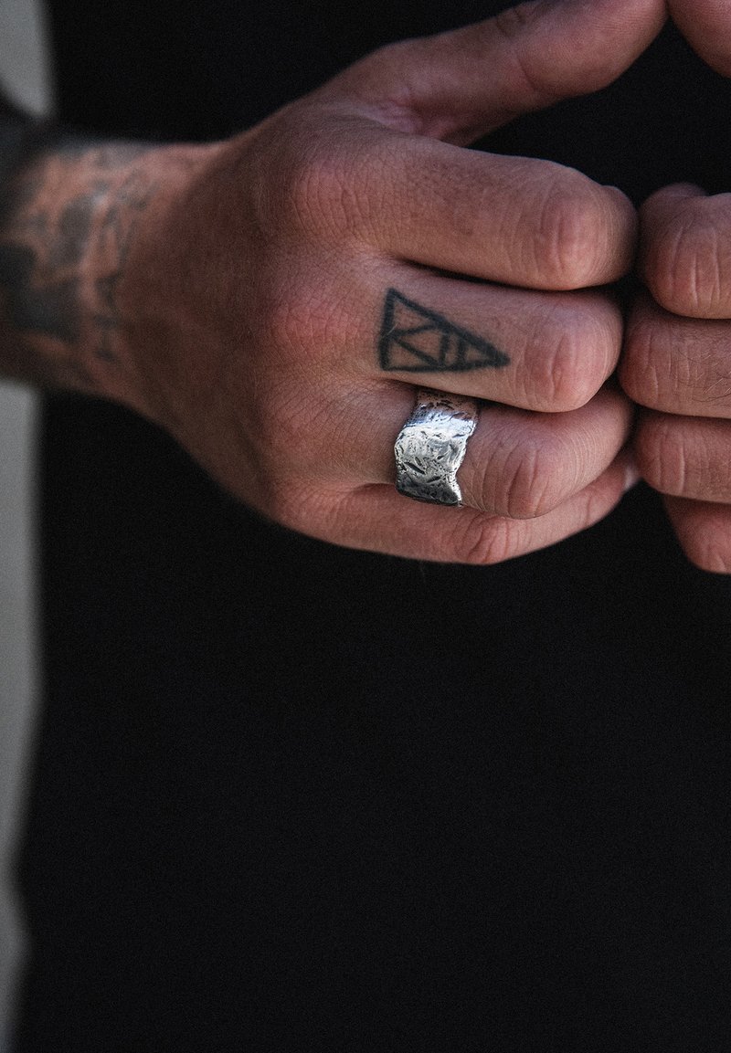 Textūruotas sidabrinis žiedas su nelygiu paviršiumi, uždedamas ant rankos su geometriniu tatuiruotė. Ranka yra atsipalaidavusi, puikiai parodanti žiedą.