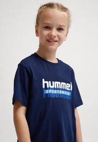Mørkeblå bomulds t-shirt med et stort hvidt og blåt "hummel SPORTSWEAR" logo, korte ærmer og rund hals.