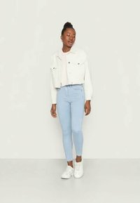 Jaqueta de denim branca cropped sobre um top rosa claro, combinada com jeans skinny azul claros e ténis brancos, de pé contra um fundo neutro.