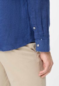 Blauwe linnen shirt met lange mouwen, knoopmanchetten en een subtiele textuur. Gecombineerd met lichtbeige broek, die een relaxte pasvorm toont.