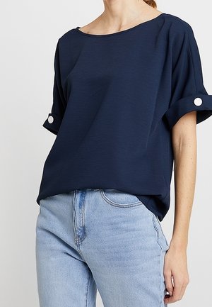 Kvinde iført en mørkeblå kortærmet top med knapmanchetter og lyseblå højtaljede jeans, stående foran en ensfarvet baggrund.