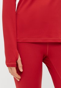 Červený top s dlhým rukávom a priliehavým strihom, so švami bez švov na manžetách, spárovaný s červenými legínami s hladkou textúrou a minimálnym šitím.