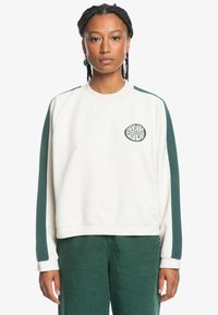 Sudadera blanca de forro polar con detalles y logo en verde, cuello redondo y puños de canalé. Combinada con pantalones de pana verde.