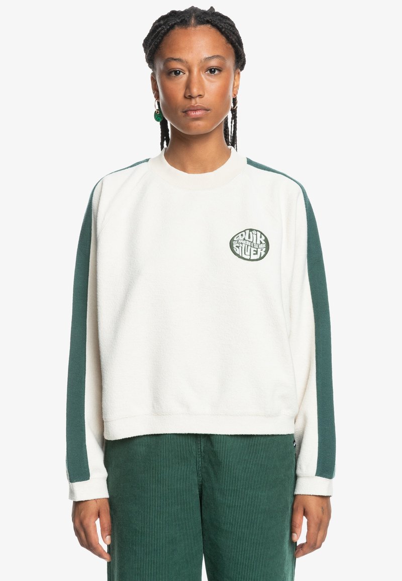 Sudadera blanca de forro polar con detalles y logo en verde, cuello redondo y puños de canalé. Combinada con pantalones de pana verde.