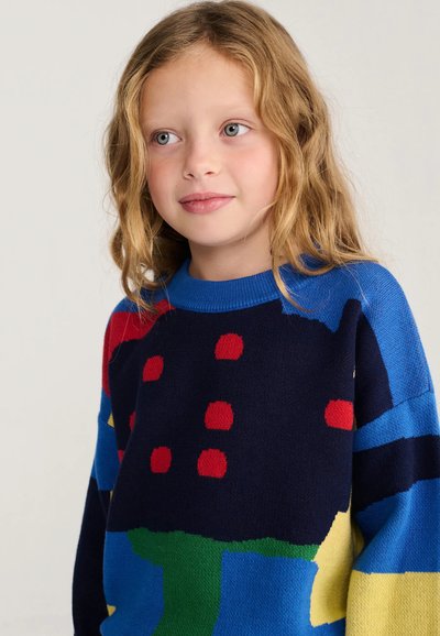 Jeune fille aux cheveux blonds ondulés portant un pull coloré avec des motifs abstraits bleus, noirs, rouges, verts et jaunes sur un fond neutre.