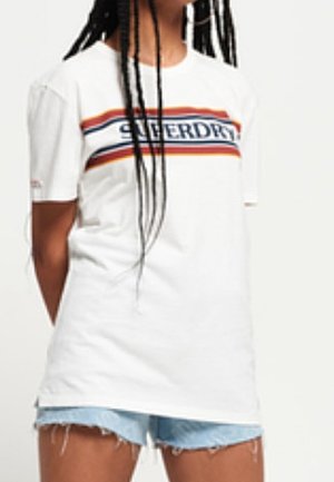 Person iført hvid Superdry t-shirt med røde og blå striber og lyseblå denimshorts med frynsete kanter.
