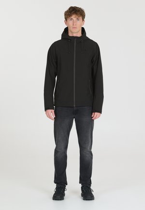 Veste noire à capuche avec zip sur le devant, poignets élastiques et poches latérales. Portée avec un jean foncé et des bottes de randonnée noires. Texture de tissu lisse.