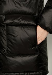 Veste doudoune noire avec une texture matelassée confortable, un grand poche extérieure et une finition lisse et brillante.