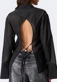 Chemise noire transparente à manches longues avec col et détail dos ouvert maintenu par des lanières nouées. Tissu texturé et coupe décontractée.