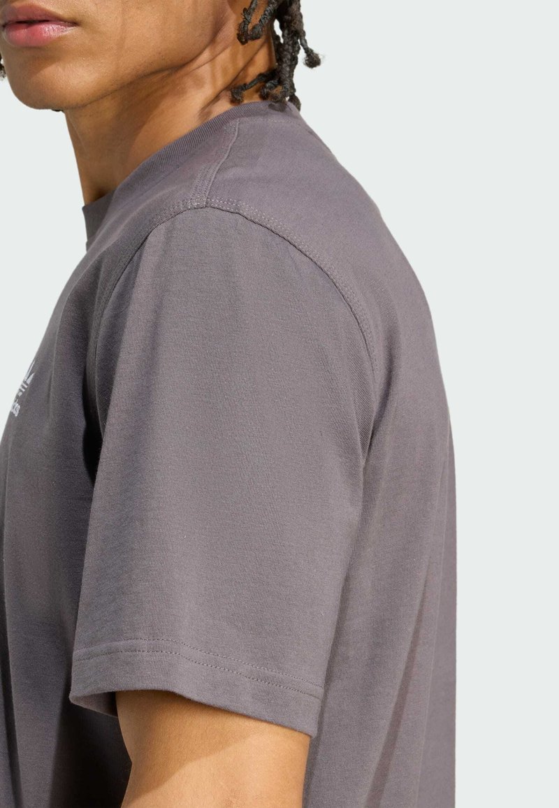 Vista laterale di una persona con capelli intrecciati che indossa una maglietta grigia a maniche corte semplice su uno sfondo chiaro.