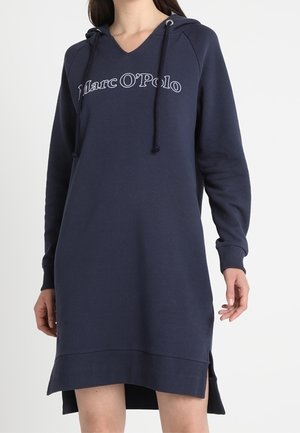 Femme portant une robe à capuche à manches longues bleu marine avec le logo "Marc O'Polo" et des fentes latérales, debout devant un fond blanc uni.