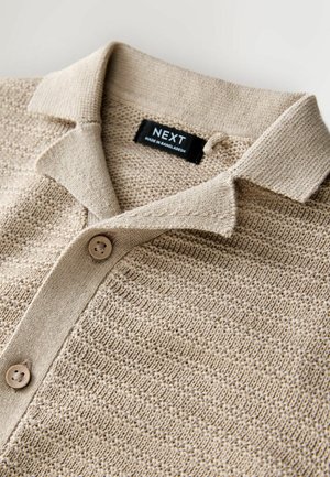 Beige strikket polo-shirt med et tekstureret mønster, en krave og to knapper. Etiket med teksten "NEXT, Made in Bangladesh." Nærbillede.