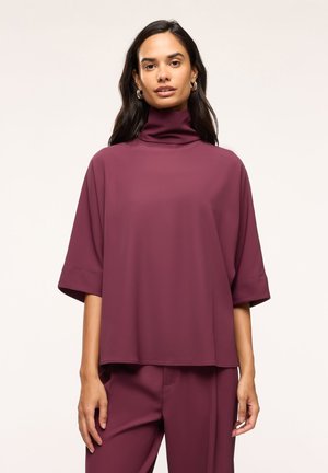 Donna che indossa una maglia bordeaux larga, a maniche corte e collo alto, e pantaloni abbinati, in piedi davanti a uno sfondo chiaro e uniforme.