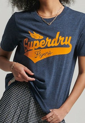 Kvinde iført en marineblå Superdry Flyers t-shirt med orange tekst og et ternet skørt, der holder i skjortens kant.