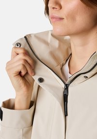 Giacca impermeabile beige con colletto alto, zip nera e bottoni a pressione. Tessuto liscio, con una fodera interna bianca e dettaglio del logo.
