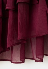 Tissu transparent superposé bordeaux avec un tissage texturé et des ourlets cousus, suspendu avec des plis doux et la lumière passant à travers les couches inférieures.