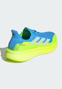 Zapatilla de running Adidas Ultraboost en azul y amarillo neón, con rayas blancas y amortiguación lightboost, sobre un fondo sencillo.
