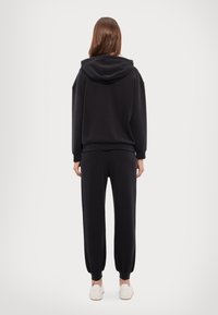 Felpa nera con cappuccio e pantaloni jogger abbinati in tessuto morbido. Presenta un cappuccio con coulisse, un taglio morbido, polsini a costine e orli alle caviglie. Sneakers bianche visibili.